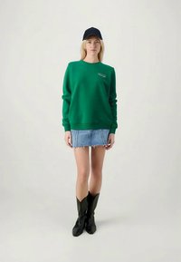 Green sweatshirt with ribbed crew neck and cuffs, paired with a denim mini skirt and black knee-high boots. Blue cap completes the outfit.