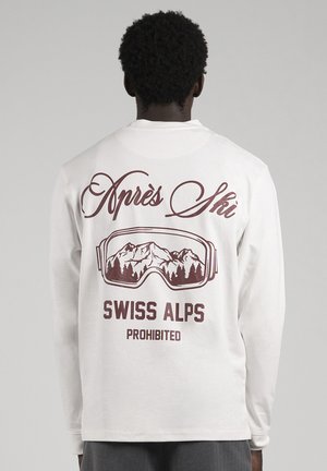 Une personne vue de dos porte une chemise blanche à manches longues imprimée avec « Après Ski », des lunettes de ski, des sommets montagneux, « Swiss Alps » et « Prohibited ».