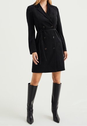 Femme portant une robe blazer noire en velours côtelé à double boutonnage avec une ceinture à la taille et des bottes en cuir noires montant jusqu'aux genoux.