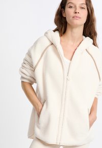 Felpa con zip in crema chiaro, dotata di cappuccio, tasche laterali e rifiniture a contrasto lungo le cuciture. Materiale morbido e texturizzato.