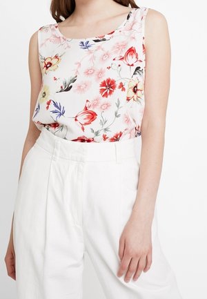 Biała bezrękawnikowa bluzka z czerwonym, różowym, żółtym i fioletowym kwiatowym wzorem, wpuszczona w białe, wysokiego stanu spodnie.