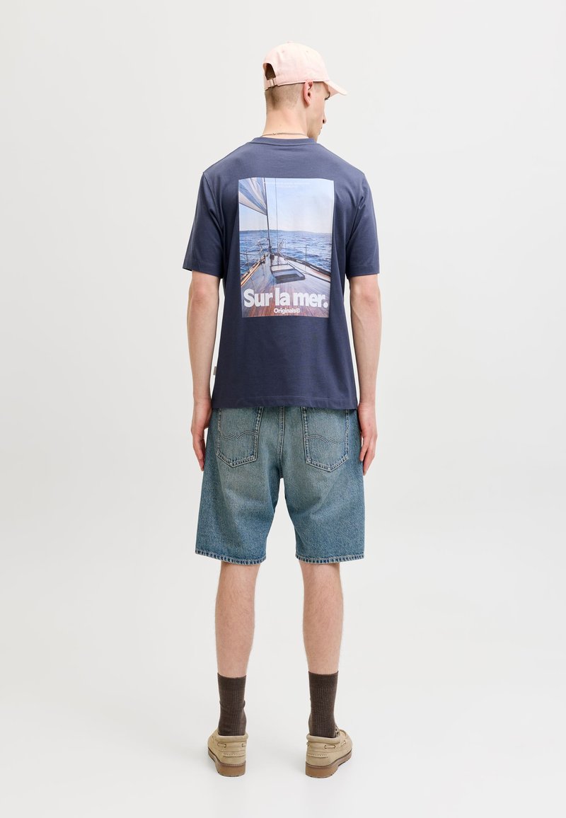 Άνδρας που φοράει navy t-shirt με εκτύπωση ιστιοφόρου και θάλασσας και κείμενο, τζιν σορτς, μπεζ παπούτσια, καφέ κάλτσες και ανοιχτό ροζ καπέλο γυρίζοντας στο πλάι.