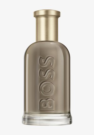 Høy, rund glassparfymeflaske med matt gulllokk og forhøyet "BOSS"-tekst på en røykgrå gjennomsiktig kropp.
