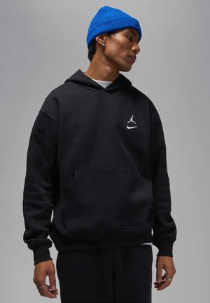 Svart bomullshuvtröja med framficka, prydd med vita Nike- och Jordan-logotyper. Bärs med en blå stickad mössa och svarta byxor.