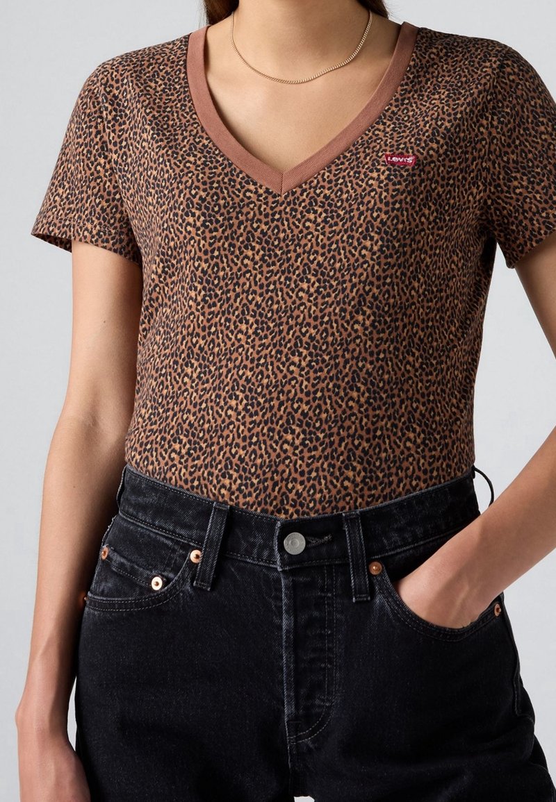 T-shirt à manches courtes col V imprimé léopard avec petit logo rouge, associé à un jean noir taille haute, main dans la poche et collier fin en or.