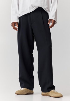 Bărbat purtând pantaloni largi negri, adidași bej și o cămașă albă cu mâneci lungi, pe un fundal simplu.