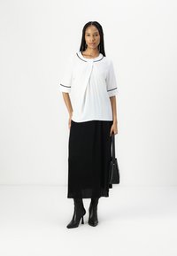 Blouse blanche avec un col arrondi et des passepoils noirs, associée à une longue jupe plissée noire. Le modèle tient un sac à main noir et porte des bottes à talons.