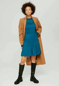 Mulher com cabelo Cacheado a usar um vestido azul, um casaco de camelo e botas altas pretas com atacadores, de pé com uma mão no bolso.