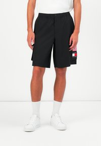 Pantaloni scurți negri cu talie elastică, buzunare laterale și o etichetă decorativă cu un steag. Material neted, design până la genunchi.