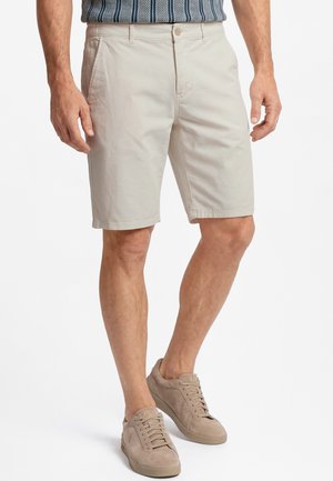 Mann trägt beige knielange Shorts, gestreiftes, in die Hose gestecktes Hemd und beige Schnürsneaker und steht vor weißem Hintergrund.