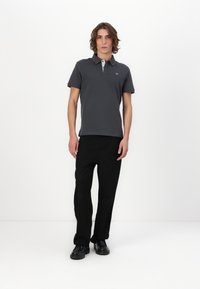Tricou polo gri închis cu guler contrastant, asortat cu pantaloni negri și pantofi negri, pe un model, pe un fundal alb.