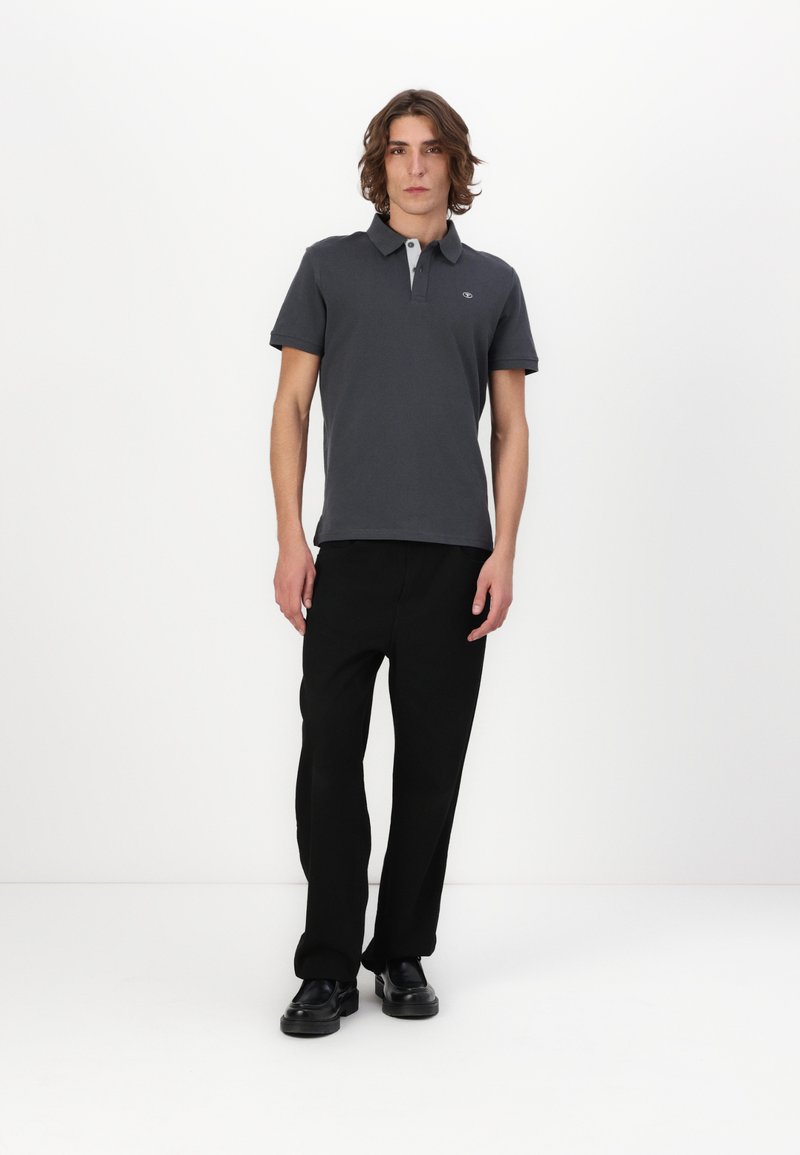Tricou polo gri închis cu guler contrastant, asortat cu pantaloni negri și pantofi negri, pe un model, pe un fundal alb.