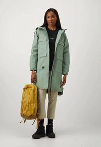 Egy nő világoszöld kapucnis kabátot visel, fekete pólót, bézs nadrágot és fekete csizmát, mellette sárga hátizsákot tart.