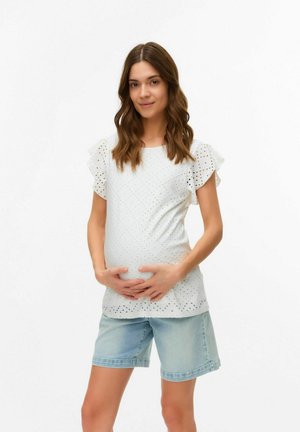Blouse blanche avec des détails en broderie et des manches volantes, associée à un short en denim bleu clair. Le modèle pose avec les mains sur son ventre.