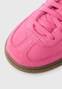 Ροζ πάνινο παπούτσι από σουέντ με υφασμένο πάνω μέρος, επίπεδα κορδόνια και καφέ λαστιχένια σόλα με τριγωνικό σχέδιο στην άκρη.