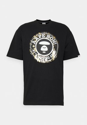 T-shirt noir avec un grand logo circulaire doré et blanc représentant un visage de singe stylisé et le texte "APE NOW ! BATHING APE."