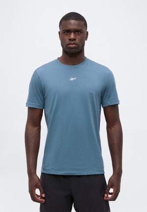 Ung mand iført en blå kortærmet T-shirt med et lille hvidt logo på brystet og sorte shorts, stående mod en ensartet baggrund.