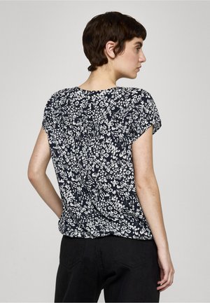 Žena s krátkými tmavými vlasy, oblečená v černo-bílé krátkosleevé květované blůze a černých kalhotách, stojící bokem na jednobarevném pozadí.