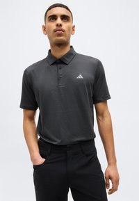 Νέος άνδρας που φοράει μαύρο πλεκτό κοντομάνικο πόλο πουκάμισο και μαύρο παντελόνι, στέκεται με το ένα χέρι στην τσέπη του, κοιτάζοντας ελαφρώς προς τα πάνω.