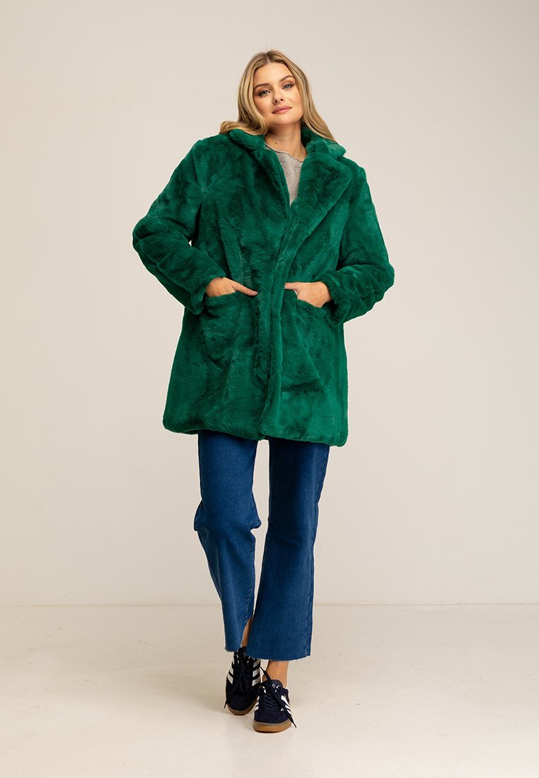 Cappotto di pelliccia sintetica verde con colletto a vista, tasche frontali e texture morbida, abbinato a jeans svasati blu e scarpe scure.