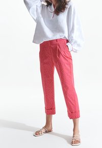 Kvinna klädd i en vit långärmad blus, rosa mönstrade kapriströjor med manschetter och vita sandaler med remmar, stående på ett vitt golv.