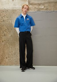 Modrá košile s krátkým rukávem a zipem s logem Nike, černé kalhoty s volným střihem a černé tenisky. Texturované pozadí se zděnou zdí.