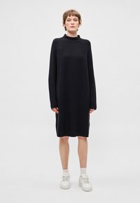 Černé pletené šaty s vysokým výstřihem, dlouhými rukávy, volným střihem a bočními průstřihy, spárované s bílými teniskami. Hladká textura.