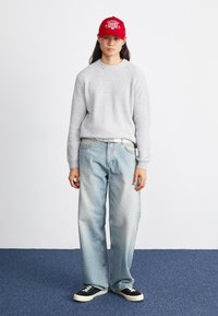 Sweater cinza com textura de nervuras, calças baggy azuis claras, ténis pretos e um boné vermelho com letras brancas, a posição em pé sobre um tapete azul contra uma parede branca.