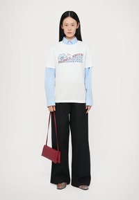 Balts T-krekls ar sarkano un zilo "Paul Smith" logo, kas kārtots virs gaiši zilā krekla ar apkakli. Apvienots ar melniem plašiem biksēm un sarkanu rokassomu.