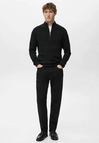 Czarny prążkowany sweter z półzamkiem przy dekolcie, zestawiony z czarnymi spodniami o prostych nogawkach i czarnymi butami, na neutralnym tle.
