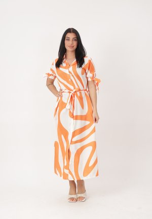 Mujer con cabello largo y oscuro que lleva un vestido midi abotonado con estampado naranja y blanco, con cinturón en la cintura y mangas cortas, de pie.
