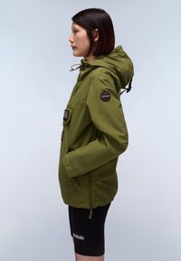 Olivegrüne Kapuzenjacke mit Reißverschluss und Patch-Applikationen. Sie hat elastische Bündchen, eine Fronttasche und eine glatte Textur. Getragen mit schwarzen Shorts.