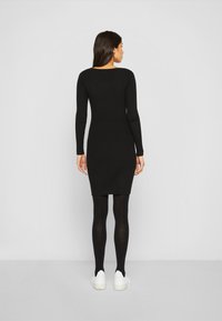 Abito lungo a maniche lunghe di colore nero con costine, silhouette aderente, che arriva sopra il ginocchio. Indossato con collant neri e sneakers bianche, su uno sfondo neutro.
