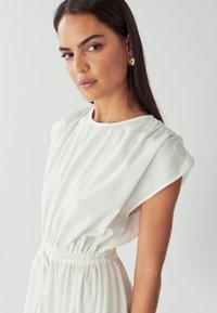 Bílé šaty s nařasenými krátkými rukávy a elastickým pasem. Hladká látka, volný střih a detail se šňůrkou v pase.