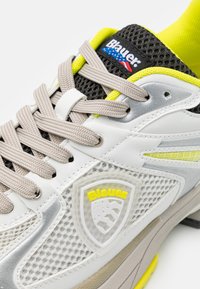 Scarpa da ginnastica bianca e grigia con texture a rete, dettagli gialli e lacci grigi. Presenta un logo a scudo e una suola rigata.