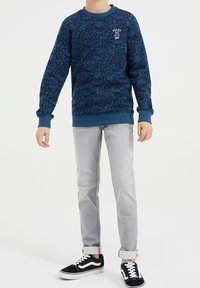 Blå sweatshirt med texturerat mönster, ribbade ärmslut och halsringning, med en logotyp på bröstet. Buren tillsammans med grå jeans och svarta sneakers.