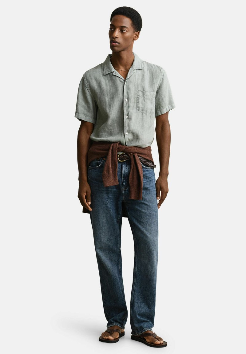 Giovane uomo che indossa una camicia a maniche corte grigio chiaro, jeans blu, maglione marrone annodato in vita, cintura e sandali marroni, in piedi contro uno sfondo semplice.