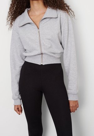 Mujer con chaqueta corta gris claro con cremallera y mangas largas y leggins negros de talle alto contra un fondo liso.