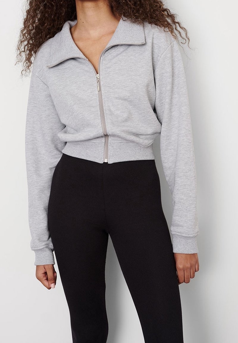 Femme portant une veste courte zippée gris clair à manches longues et des leggings noirs taille haute sur un fond uni.