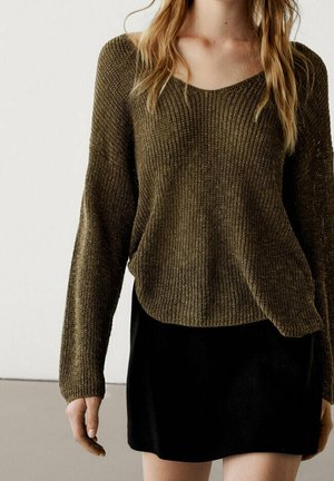 Donna che indossa un maglione lavorato a maglia largo di colore verde oliva e una gonna nera corta, in piedi contro uno sfondo chiaro e uniforme.