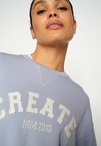 Sudadera de color azul claro con un cuello acanalado, que presenta la palabra "CREATE" y "MM2010" en un bordado blanco texturizado.