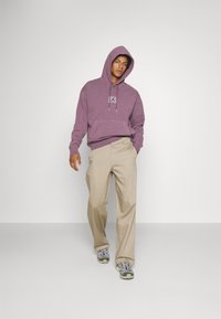 Sudadera morada con bolsillo frontal y logo en el pecho, combinada con pantalones beige y zapatillas deportivas multicolores. Diseño simple y casual.