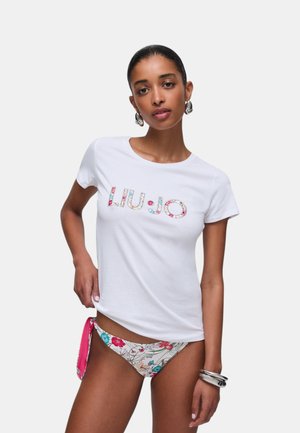 Donna che indossa una maglietta bianca con logo floreale e slip bikini floreali abbinati, in piedi con una mano sul fianco su uno sfondo semplice.