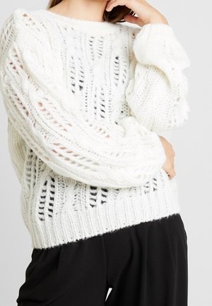 Personne portant un pull blanc en maille torsadée ample avec des manches bouffantes et un pantalon noir, posant avec une main près du visage.