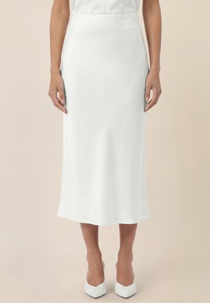Femme portant une jupe midi blanche taille haute et des talons blancs pointus, debout devant un fond beige.