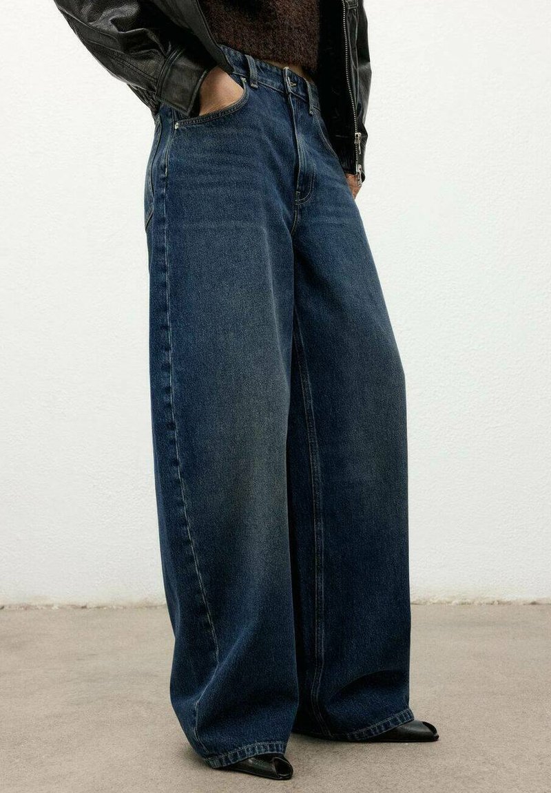Jeans de mezclilla de pierna ancha en azul oscuro, con un diseño clásico de cinco bolsillos y una textura ligeramente desgastada. Combinados con zapatos negros de punta afilada.