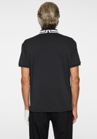 Musta poolopaita, jossa on valkoiset raidalliset kaulukset, lyhyet hihat, sileä tekstiili ja suora helma, käytetty mustien housujen ja valkoisen hansikkaan kanssa.