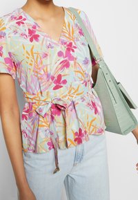Blouse peplum florale avec un décolleté en V, ornée de fleurs roses, violettes et oranges sur un fond bleu clair, nouée à la taille. Portée avec un jean clair et un sac vert.