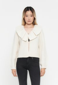 Kremfarget cardigan med en stor volangkrage, ribbede mansjetter og kant, og en frontåpning med en dekorativ knapp.