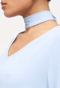 Πλαϊνή όψη ενός ατόμου που φοράει γαλάζιο μπλουζάκι με V λαιμόκοψη και έναν ταίριαζον φαρδύ υφασμάτινο κολιέ γύρω από τον λαιμό.